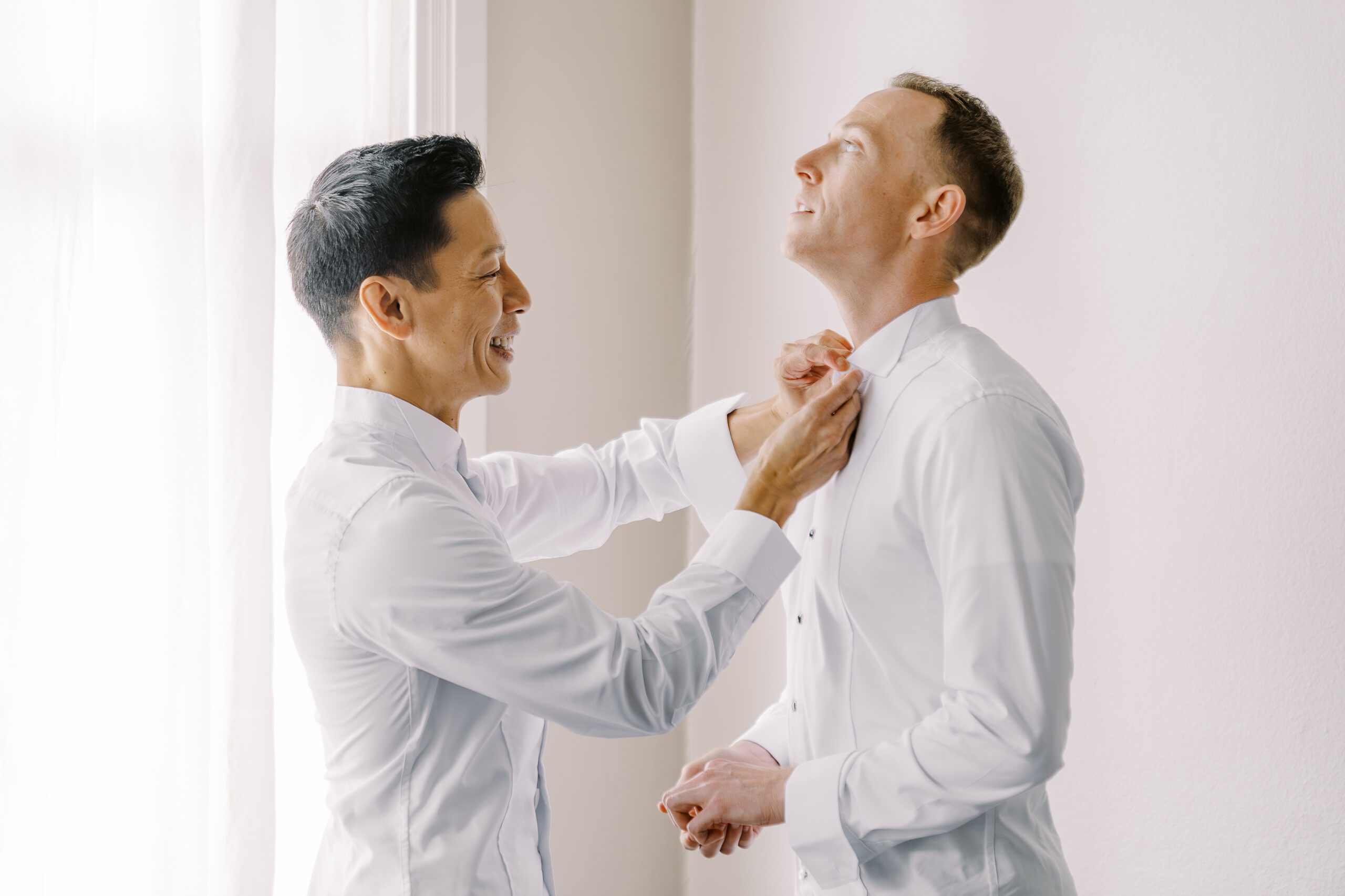 a groom helps his fiance button his shirt buttons while getting ready for their vizcaya sacramento wedding