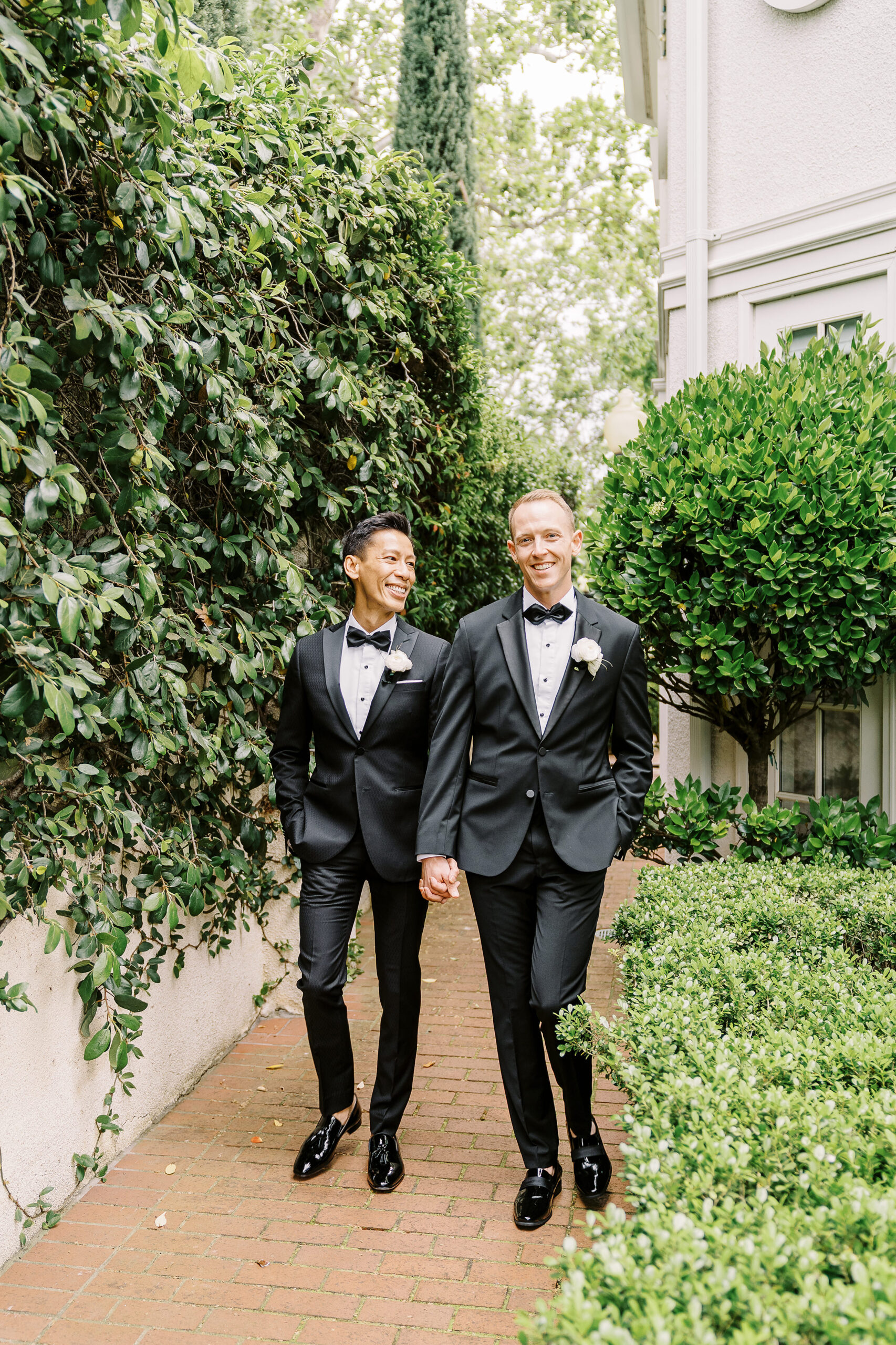 two grooms walk and laugh holding hands in front of an ivy covered wall at their vizcaya sacramento wedding