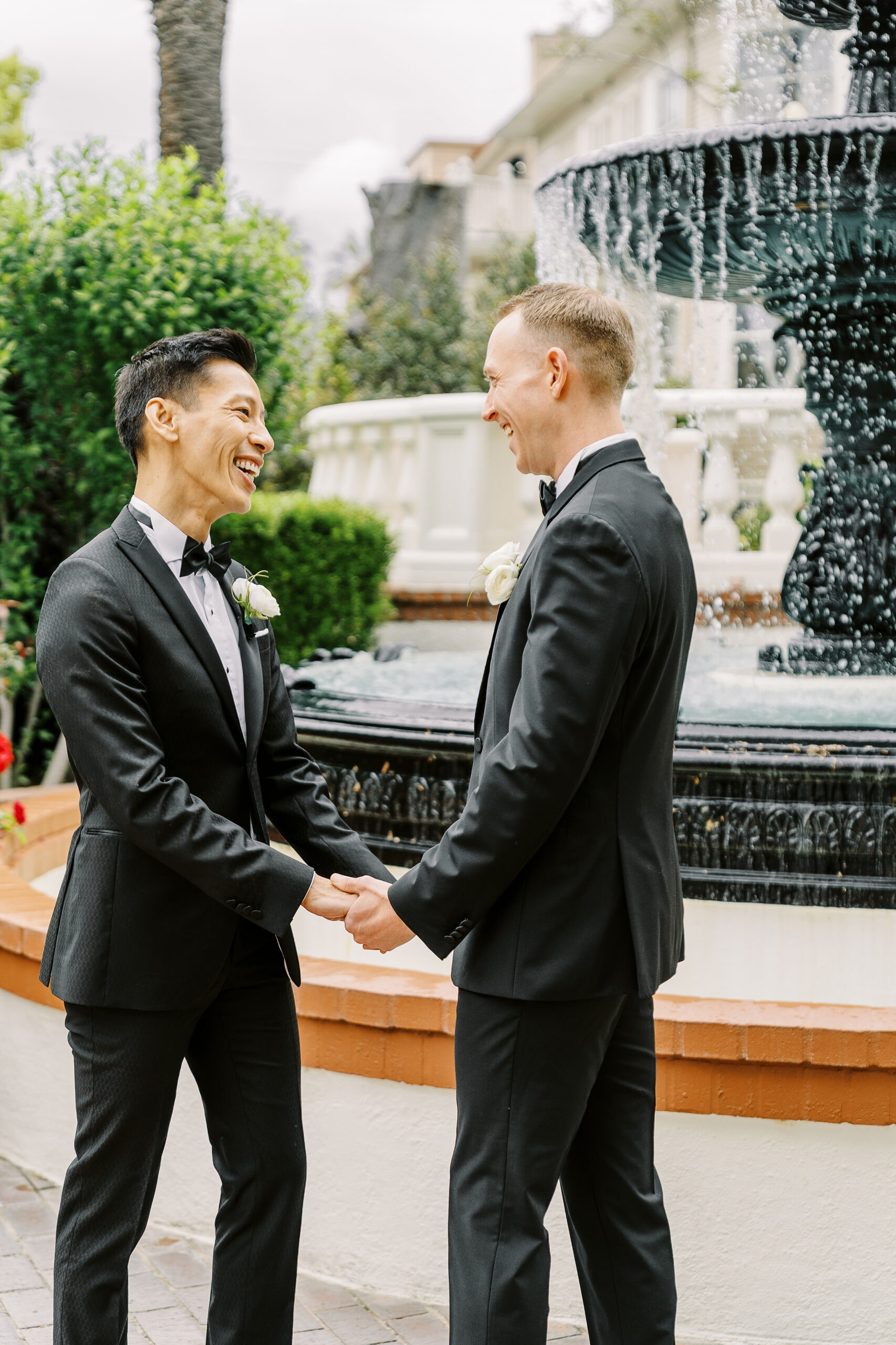 two grooms laugh together while holding hands in front of the water fountain at their vizcaya sacramento wedding