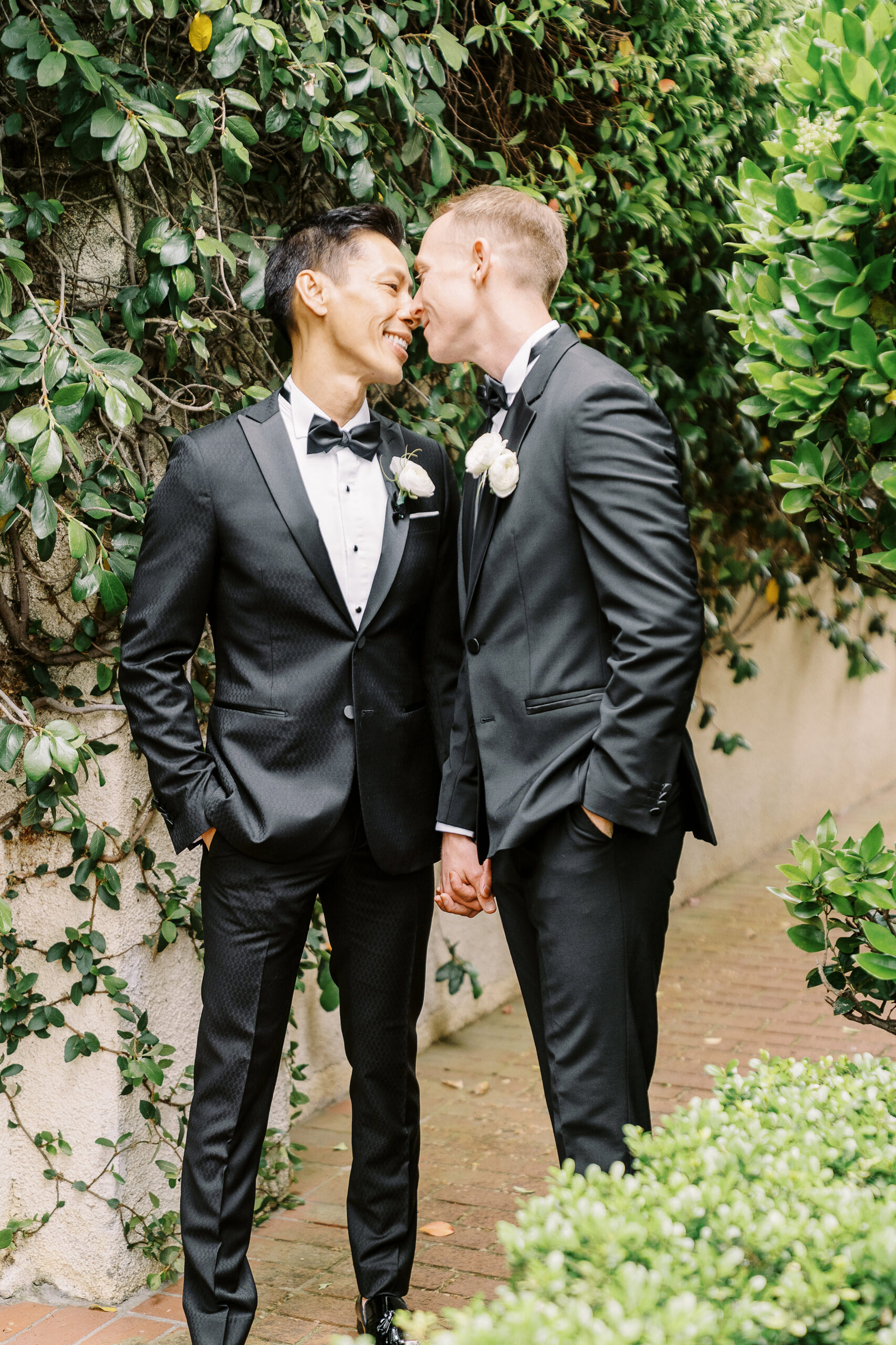 two grooms nuzzle noses while holding hands in front of an ivy covered wall at their vizcaya sacramento wedding