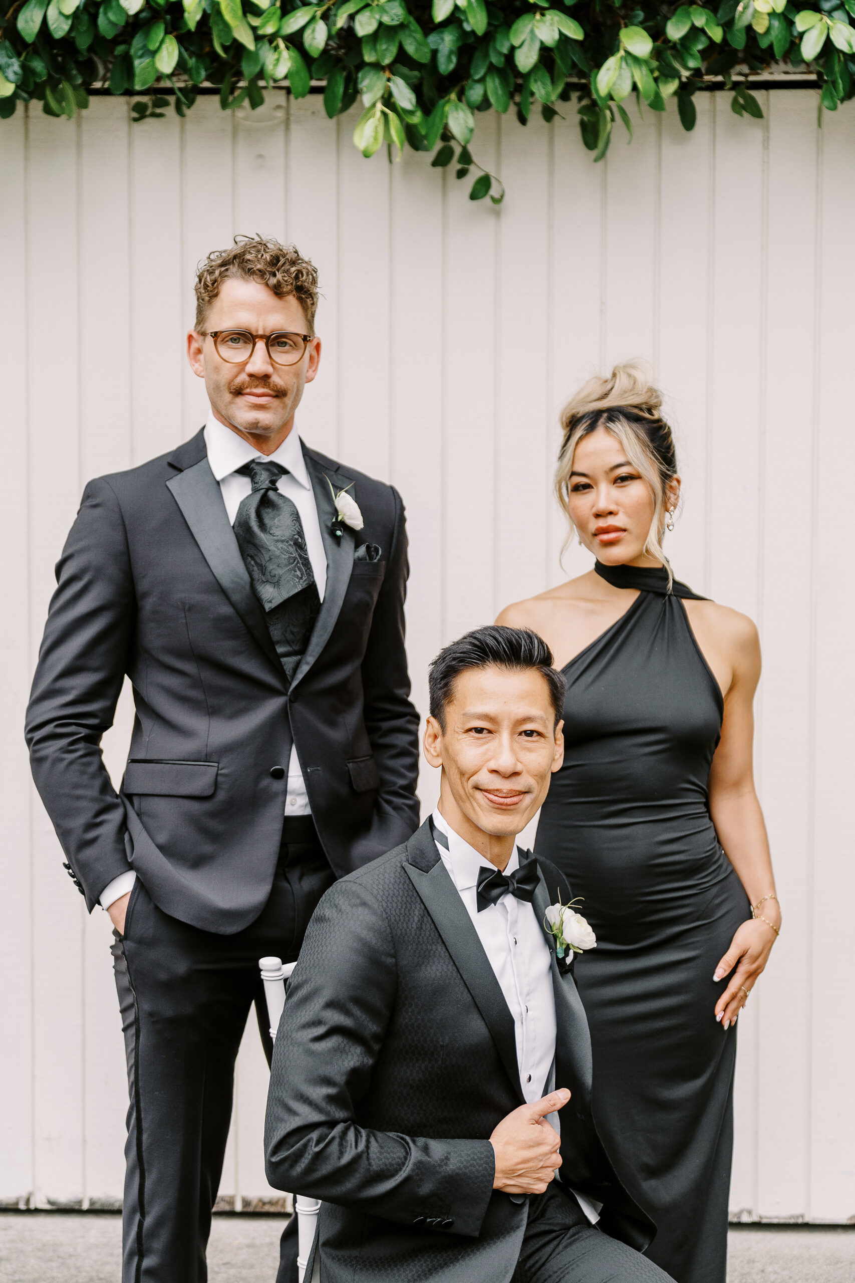 a groom poses with his wedding party members in his sleek black tux in front of a white wall