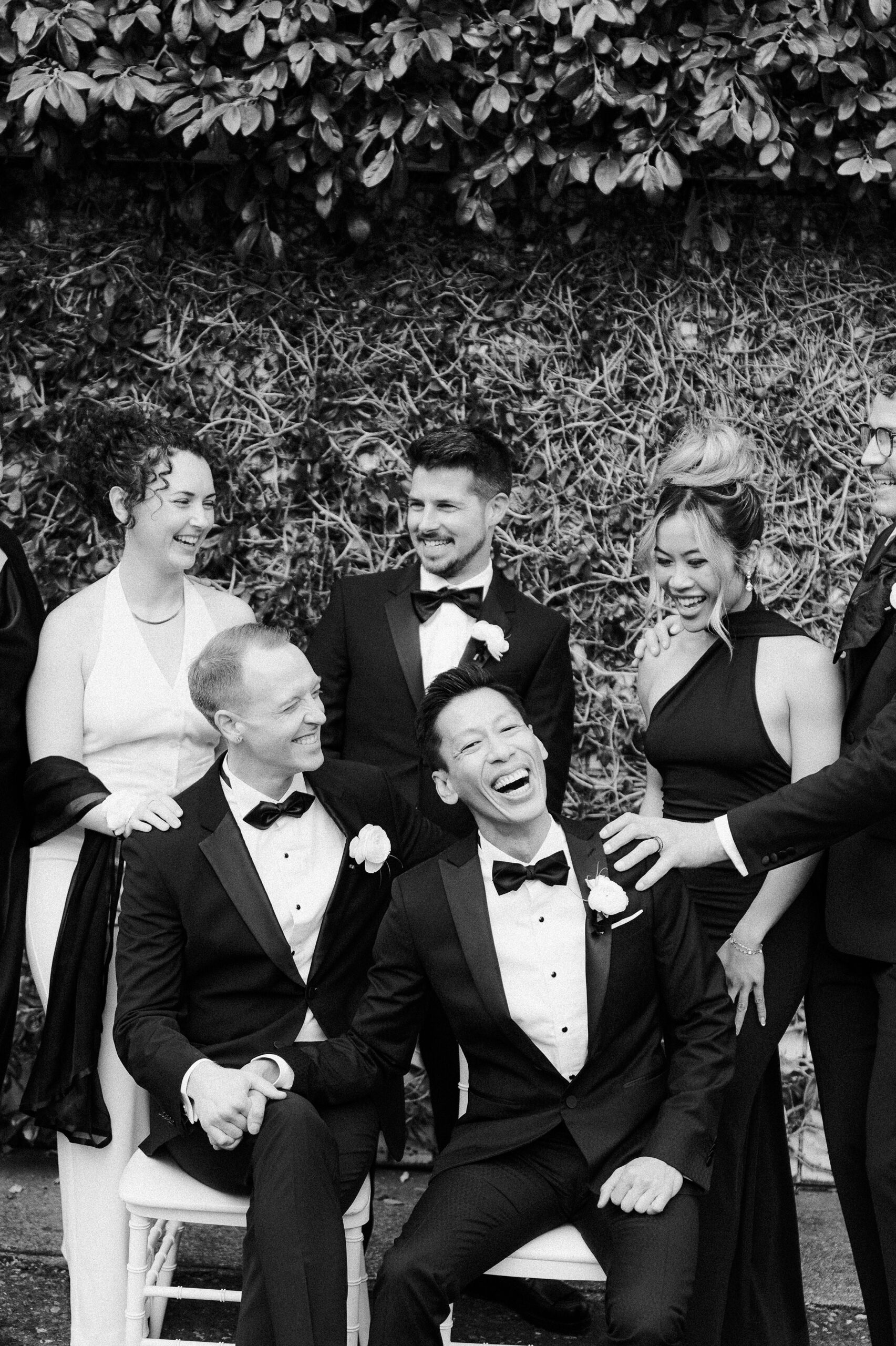 a black and white photo of two grooms laugh with their wedding party in sleek black tuxes at the vizcaya sacramento wedding venue